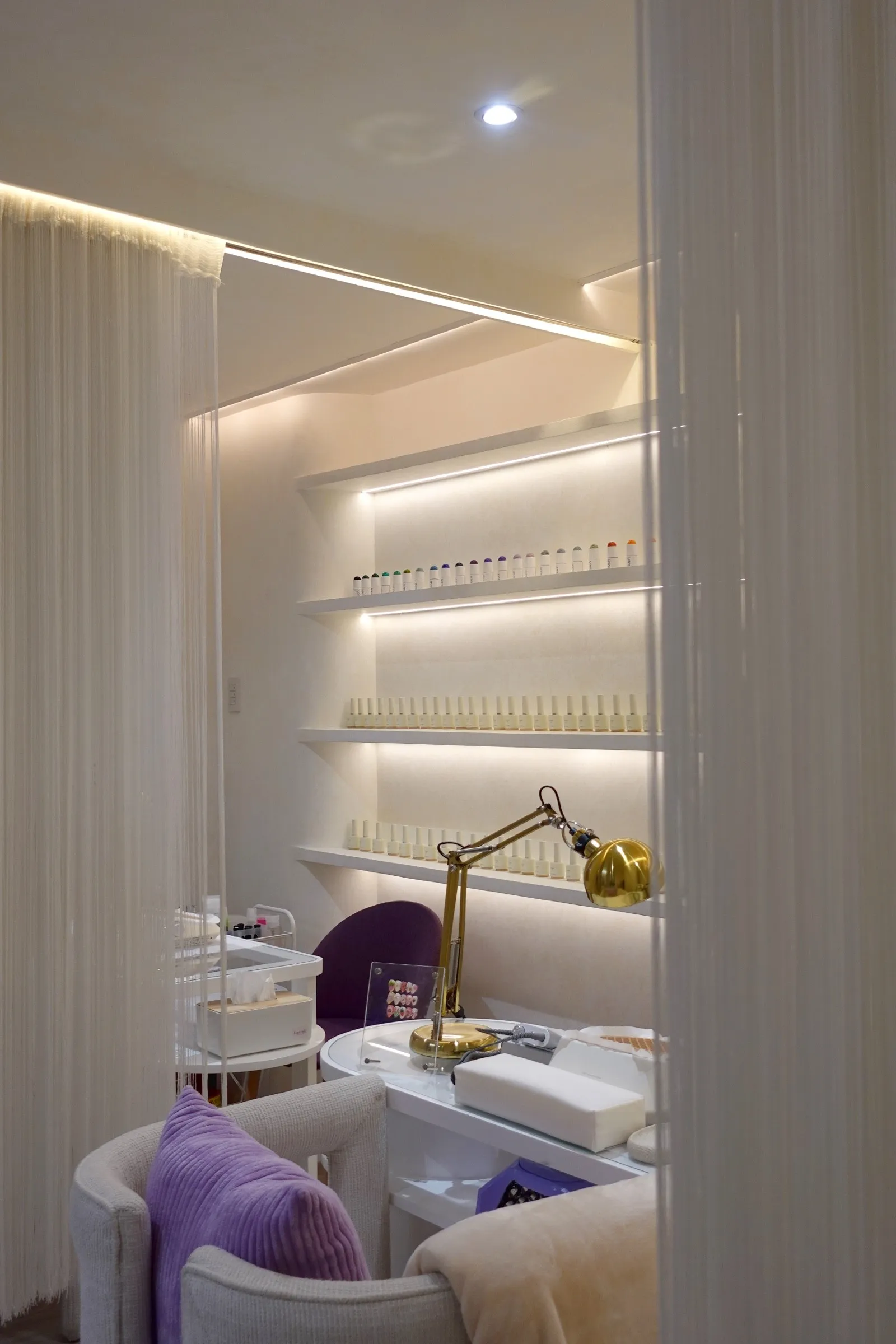 A nail station at Lavande with a brass lamp, lit shelves of polish and a lilac cushion
